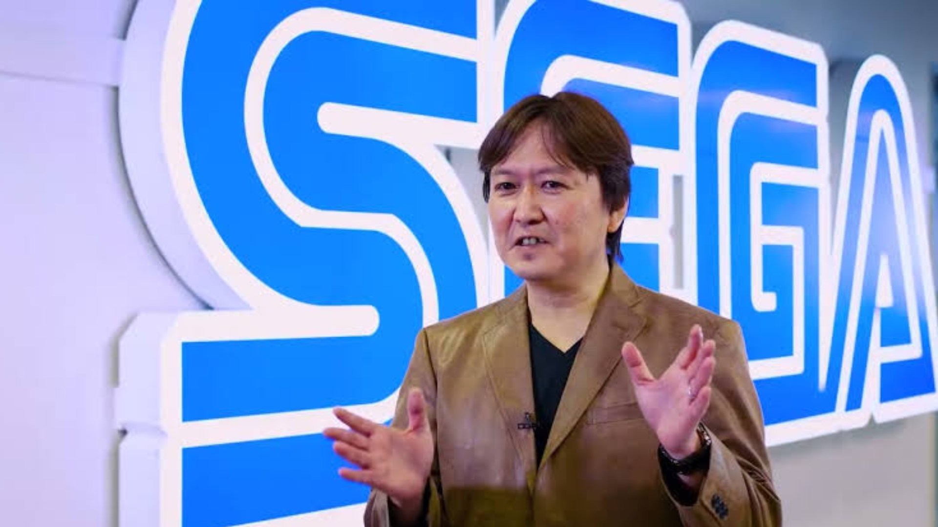 Takashi Iizuka standing in front of Sega Logo talking